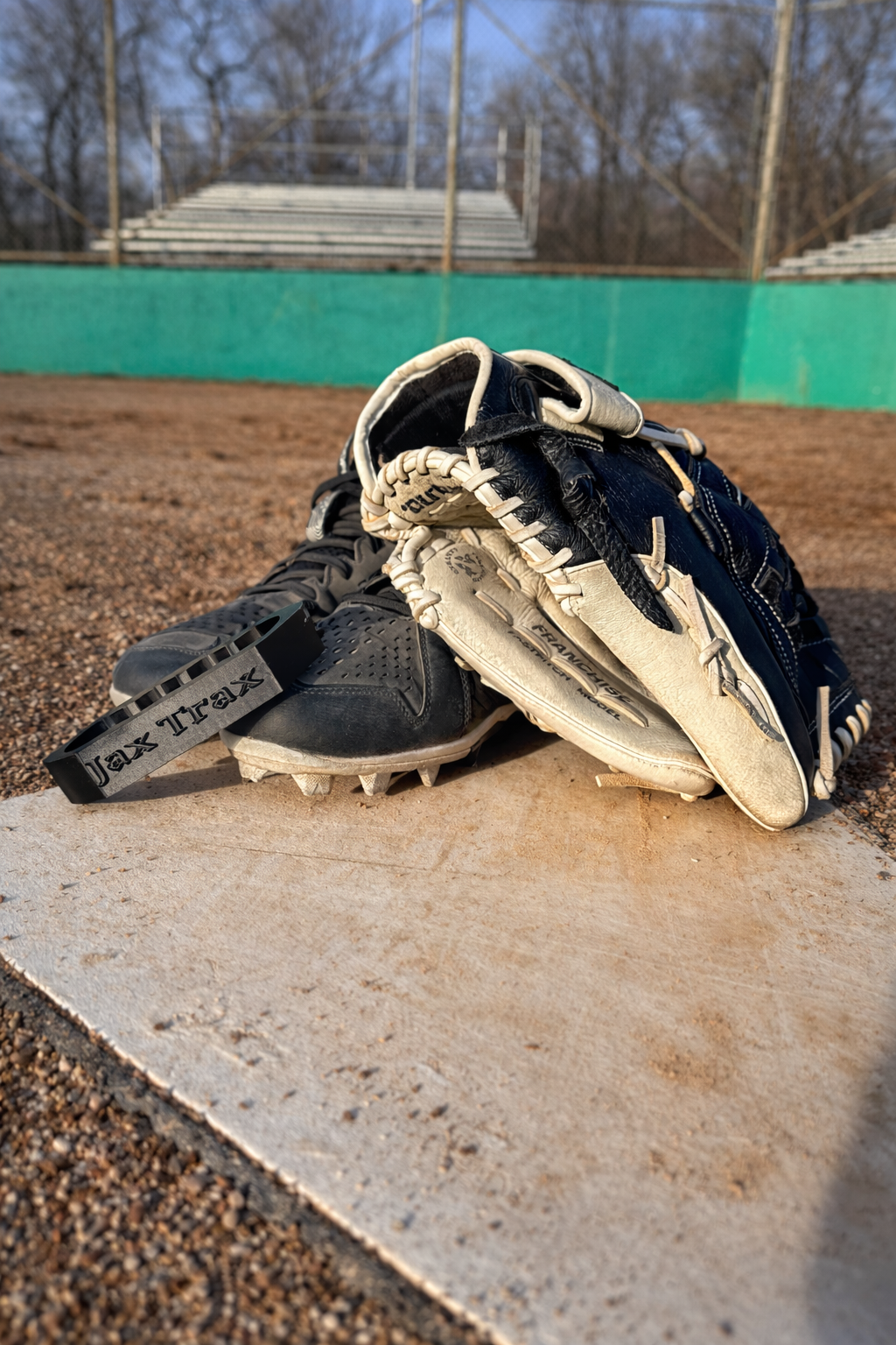 Baseball cleats and a glove resting on a base, with a black boot-cleaning tool labeled “Jax Trax” placed beside the cleats on a dirt field.