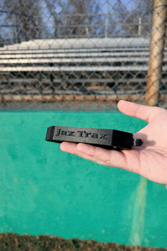 Hand holding a black boot-cleaning tool labeled “Jax Trax” in front of a green fence and empty metal bleachers.